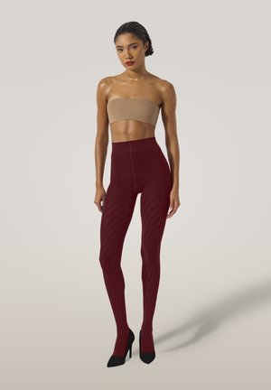 Vrouw met strak naar achteren gekamd haar, gekleed in een beige bandeau top, gemarmerde bordeauxrode hoger geplaatste legging en zwarte hoge hakken, staande tegen een lichte achtergrond.