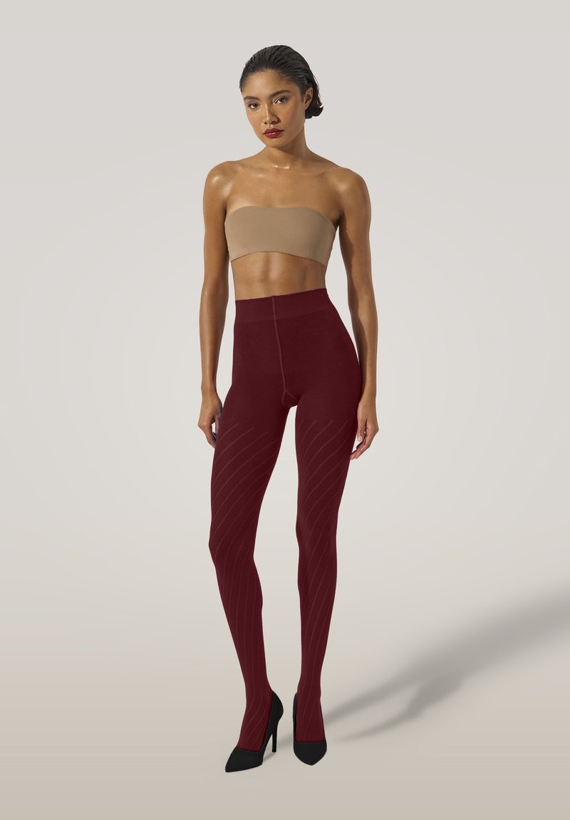 Kvinna med slickat hår iförd en beige bandeau topp, ribbade vinröda högmidjade leggings och svarta högklackade skor som står mot en ljus bakgrund.