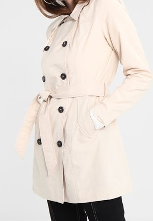 Kvinde iført en beige dobbeltradet trenchcoat med sorte knapper, taljebælte og hænder i lommerne mod en ensfarvet baggrund.
