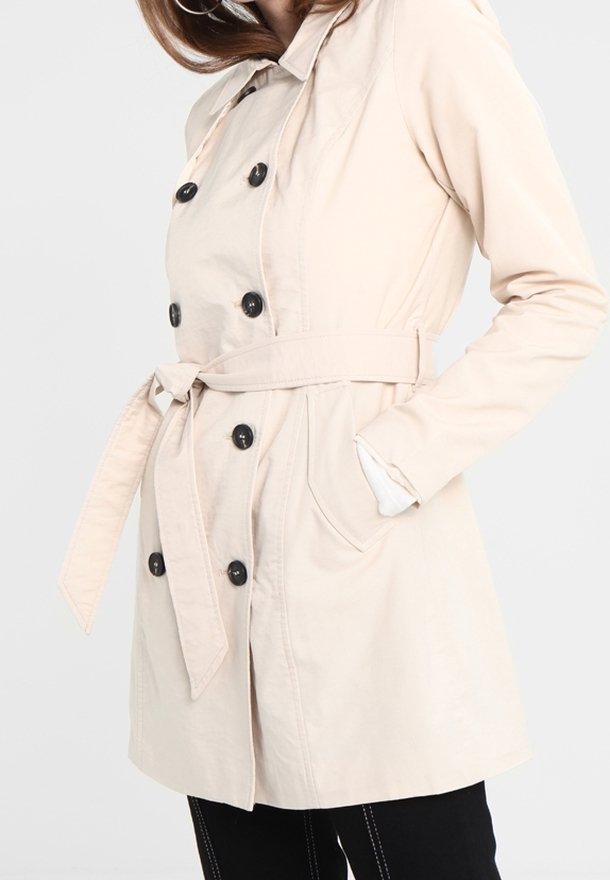 Femme portant un trench-coat beige à double boutonnage avec des boutons noirs, une ceinture à la taille et les mains dans les poches sur un fond uni.