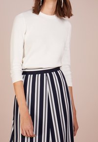 Donna che indossa un top bianco a maniche lunghe con texture e una gonna blu marina con strisce verticali bianche e nere, in piedi contro uno sfondo beige.