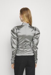 Blusa argentata in satin con maniche a sbuffo, colletto alto e dettagli arricciati sul retro. Abbinata a pantaloni neri completa il look.