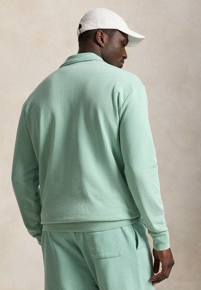 Άνδρας που φοράει ανοιχτοπράσινο μακρυμάνικο πουκάμισο και παντελόνι με λευκό καπέλο μπέιζμπολ, κοιτάζοντας πλάγια σε ουδέτερο φόντο.