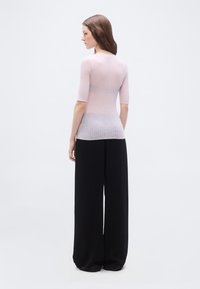 Γυναίκα που στέκεται πλαγίως φορώντας ένα ροζ ριμπ πλεκτό τοπ με μανίκια μέχρι τον αγκώνα και φαρδιά μαύρη παντελόνα, μπροστά σε ένα απλό φόντο.