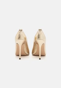 Gyldne metalliske høyhæler med spiss tå og stiletthæl. Har en dekorativ detalj bak. Glatt tekstur og slank design.