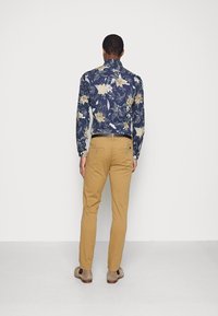 Camicia blu navy con fiori chiari, abbinata a pantaloni color cuoio e scarpe grigie. Tessuto liscio, design aderente con colletto.