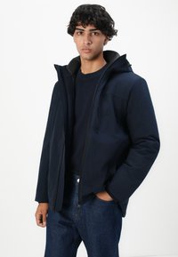 Ung mann med mørkt krøllete hår iført en marineblå hettegenser, mørk genser og jeans, stående med den ene hånden i lommen.