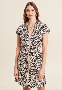Kleid aus Leopardenmuster mit kurzen Ärmeln, vorderem Reißverschluss, Bindegürtel in der Taille und zwei Brusttaschen. Cremefarbener Hintergrund mit schwarzen Flecken.