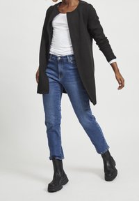 Persona che indossa jeans blu, giacca lunga nera strutturata, camicia bianca e stivali neri chunky mentre cammina su uno sfondo bianco.