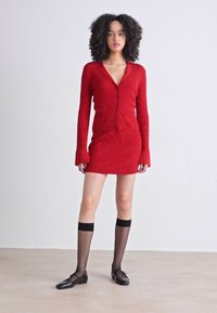 Rød ribbestrikket cardigan kombinert med en matchende mini-kjole, med lange ermer og krage. Modellen har på seg svarte thigh-high sokker og leiligheter.