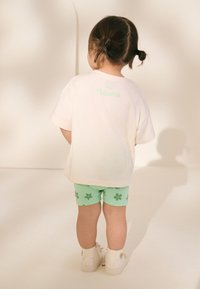 Light pink t-shirt with "Moana" printed in green, paired with mint green shorts featuring dark green floral patterns and white shoes.