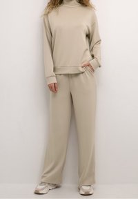 Ensemble de vêtements d'intérieur beige comprenant un haut à col roulé et un pantalon à jambes larges. Tissu doux, coupe décontractée, et poches latérales sur le pantalon.