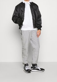 Černá saténová bomber bunda s žebrovanými manžetami, šedé lehké kalhoty a černé tenisky s bílými akcenty. Pod tím bílá mikina s kapucí.