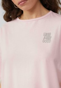 Lichtroze T-shirt gemaakt van zachte stof met een relaxte pasvorm, voorzien van een kleine grijze graphic met de tekst "IT'S ALL GOOD" op de borst.