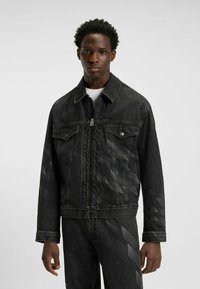 Zwarte denimjas met twee borstzakken, zichtbare stiknaden en een ritssluiting aan de voorkant. Heeft subtiele geometrische patronen op de mouwen. Soepele textuur.