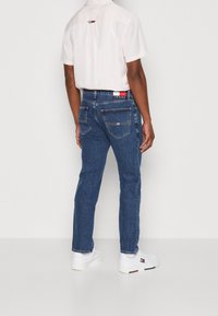 Tmavě modré džínové kalhoty s rovnými nohavicemi, s kapsami na zadní straně a štítkem s logem na pasu, v kombinaci se světlou košilí.