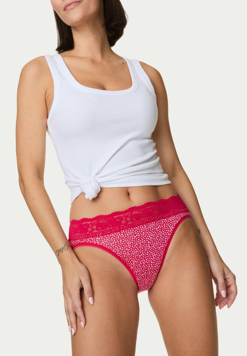 Rote Unterwäsche mit Spitzendetails und einem gemusterten Stoff, der ein gepunktetes Design aufweist. Weicher Stoff mit einer mittelhohen Schnittform. Kombiniert mit einem weißen Tanktop.