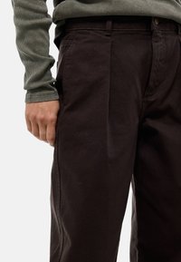 Pantalons marron foncé en tissu texturé, avec une taille haute, des poches latérales et un design à l'avant plat. Portés avec un t-shirt à manches longues côtelé vert.