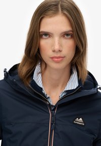 Jeune femme aux cheveux châtain clair portant une veste bleu marine avec un logo de montagne et une chemise à col rayée bleu et blanc.
