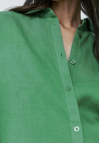 Close-up of a person wearing a green button-up shirt, focusing on the collar, buttons, and upper chest area.