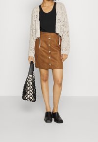 Cárdigan corto de punto de tejido abierto en color beige claro, top negro sin mangas, falda de ante marrón con botones en la parte delantera, mules negros y bolso de red negro.
