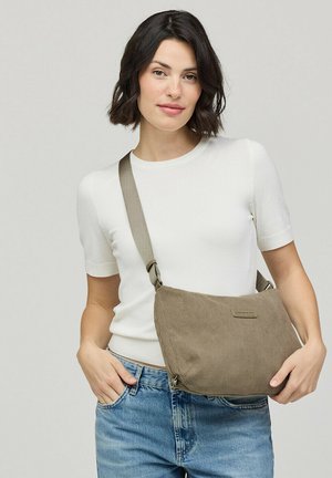 Frau mit kurzen dunklen Haaren, die ein weißes kurzärmeliges Oberteil und blaue Jeans trägt, mit einer beigen Cord-Umhängetasche.