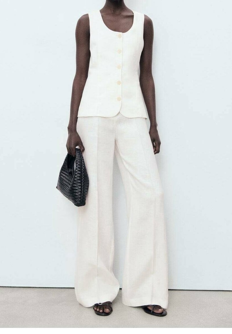 Personne à la peau foncée portant un haut blanc sans manches à boutons et un pantalon blanc à jambes larges, tenant un sac à main noir tissé et portant des sandales noires.