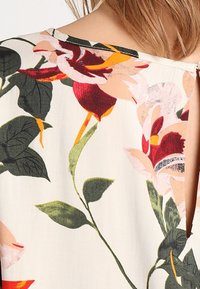 Primo piano di un tessuto beige con un grande motivo floreale in rosso, rosa, arancione e verde, che mostra un collo e parte di una spalla nuda e capelli.