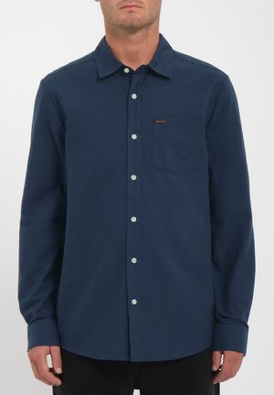 Homme portant une chemise bleu foncé boutonnée avec une poche poitrine et un col, debout devant un fond clair uni.