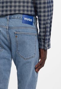 Hele sinisest teksapüksid sirge lõikega, tagataskutega ja sinise nahast sildiga, millel on valge kirjas "HUGO". Mudel kannab ruudulist särki.