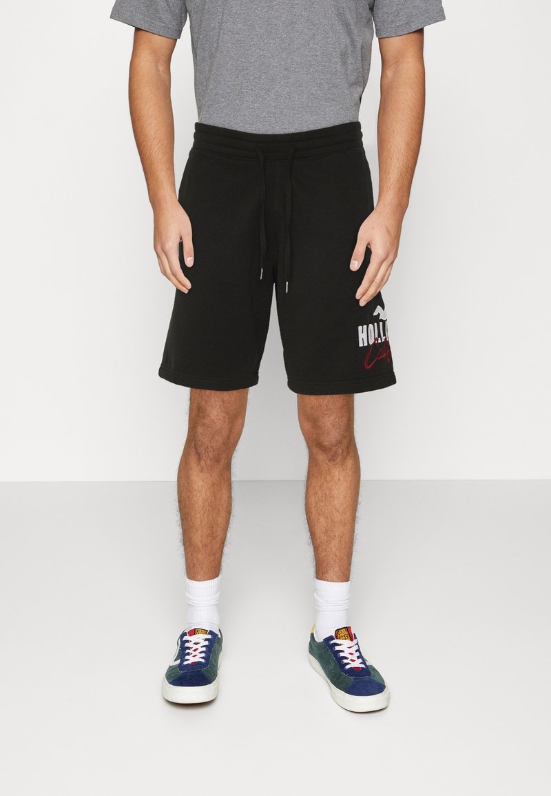 Homme portant un short noir à cordon avec un logo blanc et rouge sur la jambe, un t-shirt gris, des chaussettes blanches et des baskets bleues, debout sur un sol uni.
