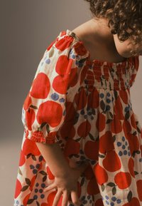 Rød og hvid kjole med puffede ærmer, prydet med et blomstermønster bestående af store røde æbler og små blå bær. Smocked bodice detalje.