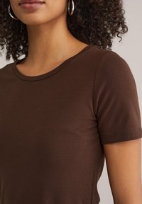 Vrouw met krullend haar die zilveren oorringen draagt en een nauwsluitend bruin shirt met korte mouwen voor een neutrale achtergrond.