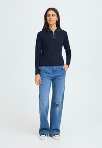 Tengeréskék bordázott pulóver fél cipzáros gallérral, világoskék, bő szárú farmernadrággal és fekete sportcipővel párosítva. Az öltözet testhez simuló vonalvezetést mutat.