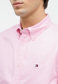 Άνδρας που φορά ρόζ και λευκό ριγωτό πουκάμισο με κουμπωτό γιακά και μικρό ορθογώνιο κόκκινο, λευκό και μπλε λογότυπο στο στήθος.