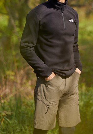 Person trägt einen schwarzen Fleece-Pullover mit Halbreißverschluss und beige Cargoshorts, die Hände in den Taschen, steht draußen vor grüner Laubkulisse.