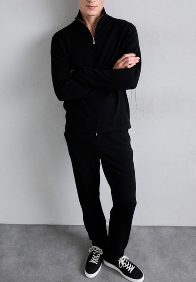 Black zip-up sweater with long sleeves, paired with matching black pants. The model wears black and white sneakers. Smooth fabric texture.