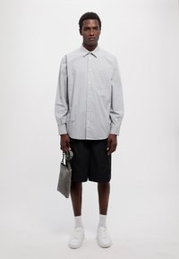 Camicia a righe con bottoni in azzurro chiaro e bianco, pantaloni corti neri, sneakers bianche, in mano una borsa grigia. Design semplice e vestibilità rilassata.