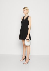 Robe noire sans manches avec un décolleté en V, jupe évasée ; associée à des talons noirs à lanières et à un petit sac à main argenté orné d'un motif texturé.