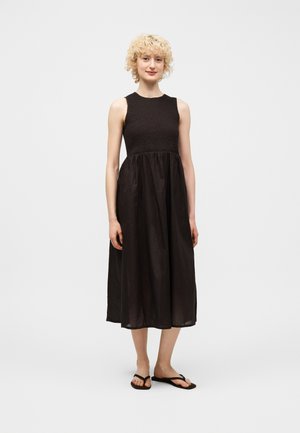 Žena s kučeravými blond vlasmi, oblečená v čiernych midi šatách bez rukávov a čiernych žabkách, stojaca pred jednoduchým svetlým pozadím.