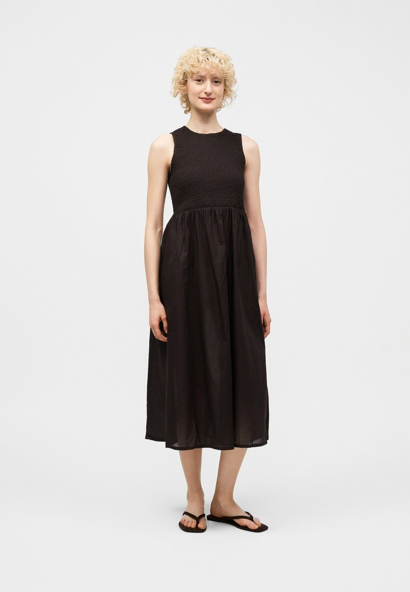 Γυναίκα με ξανθά σγουρά μαλλιά, ντυμένη με μαύρο μίντι φόρεμα χωρίς μανίκια και μαύρα σαγιονάρες, στέκεται μπροστά σε απλό ανοιχτό φόντο.