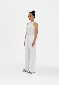 White ribbed tank top with sleeveless design, paired with elastic waistband white pants. Model stands on a plain background. Black and white shoes.