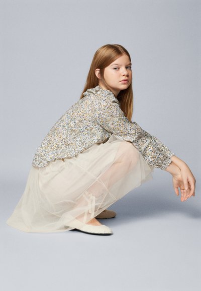 Blusa a maniche lunghe con stampa floreale e polsini volant, abbinata a una gonna di tulle avorio a strati e scarpe ballerine chiare.