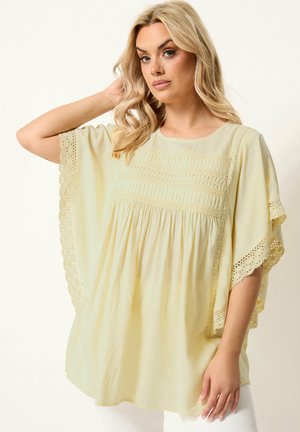 Femme blonde aux cheveux ondulés portant un chemisier ample jaune pâle avec bordure en dentelle sur les manches larges et des panneaux avant texturés.
