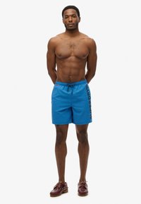 Shorts de bain bleus avec une taille à cordon et "SUPERDRY" imprimé en noir sur le côté gauche. Texture lisse, adaptés aux activités aquatiques.
