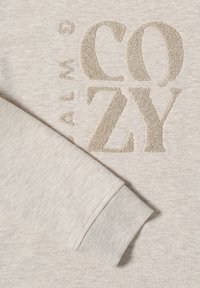 Světle béžová mikina s texturovaným zlatým nápisem "COZY". Disponuje žebrovaným manžetovým zakončením v mírně tmavším béžovém odstínu, které ukazuje jemnou texturu látky.