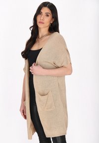 Cardigan beige in maglia con maniche corte, apertura frontale e due tasche davanti. Realizzato in un tessuto morbido e testurizzato.