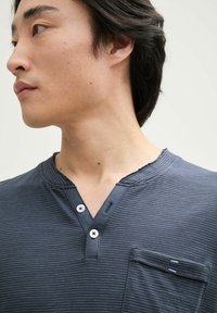 Námorně modré pruhované henley tričko s výstřihem do V, dvěma knoflíky a náprsní kapsou. Měkká látka s žebrovanou texturou a roztrženým detailem na výstřihu.