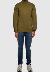 Olivenfarbener Rollkragenpullover mit gerippten Bündchen und Saum, kombiniert mit blauen Slim-Fit-Jeans und beigen Sportschuhen.