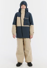 Wasserabweisende Jacke in Marineblau und Beige mit frontaler Reißverschluss, einer Brusttasche und Kapuze. Kombination mit locker sitzenden Beigen Hosen und schwarzen Stiefeln.